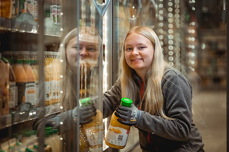 En elev från försäljnings- och serviceprogrammet ler och ställer in en flaska juice i en kyldisk på ICA Kvantum under sin lärlingsplats.